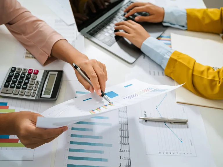 Two pairs of hands working on documents with charts, a computer in the top left corner and a calculator on the righthand side are also visible